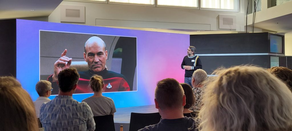 Foto von Lars Müller bei seinem Ignite Talk auf der Digitalen Woche Kiel. Seine Präsentation zeigt ein Foto von Jean-Luc Picard auf dem Raumschiff Enterprise.